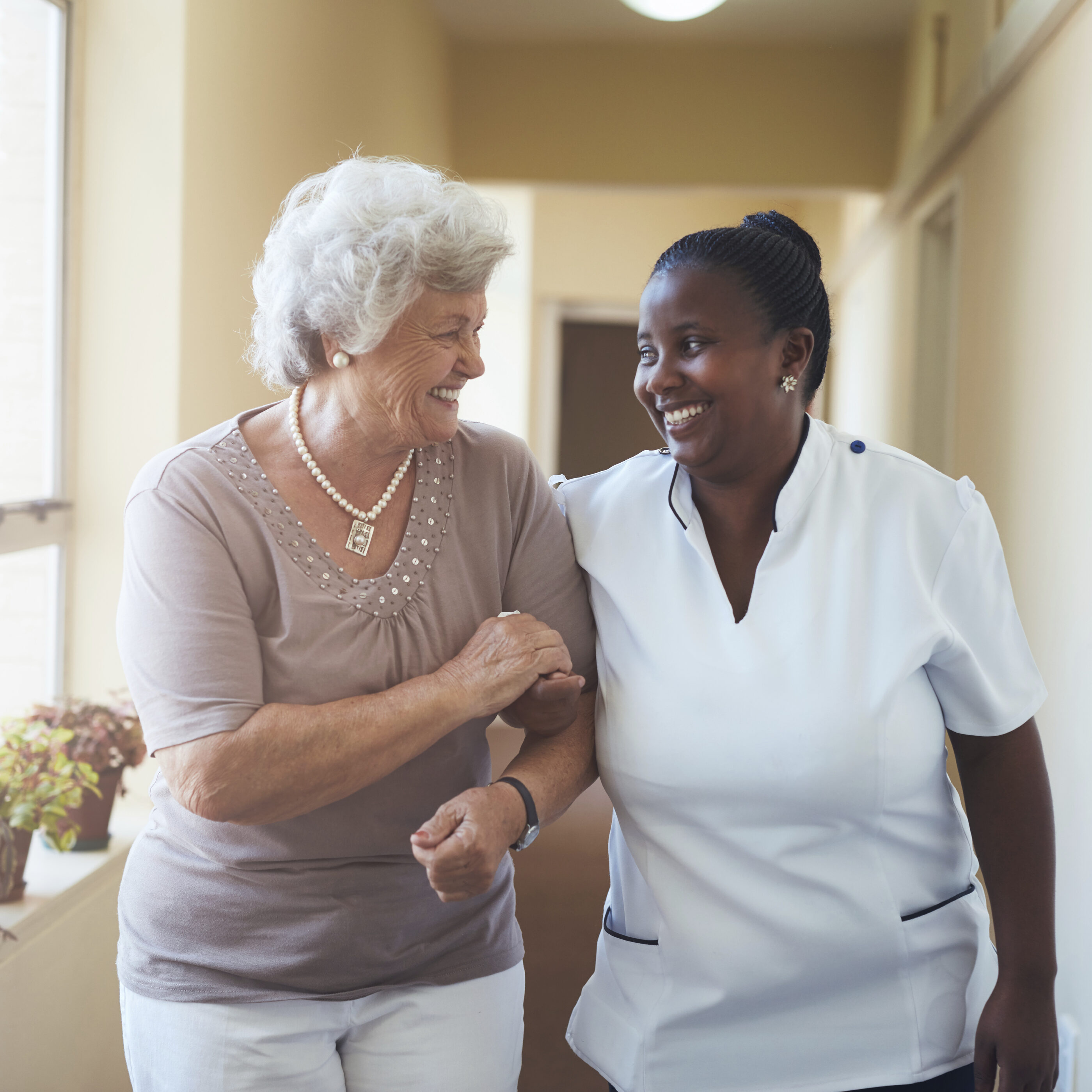 Portrait of smiling home caregiver and senior woman walking together through a corridor. Healthcare worker taking care of elderly woman.