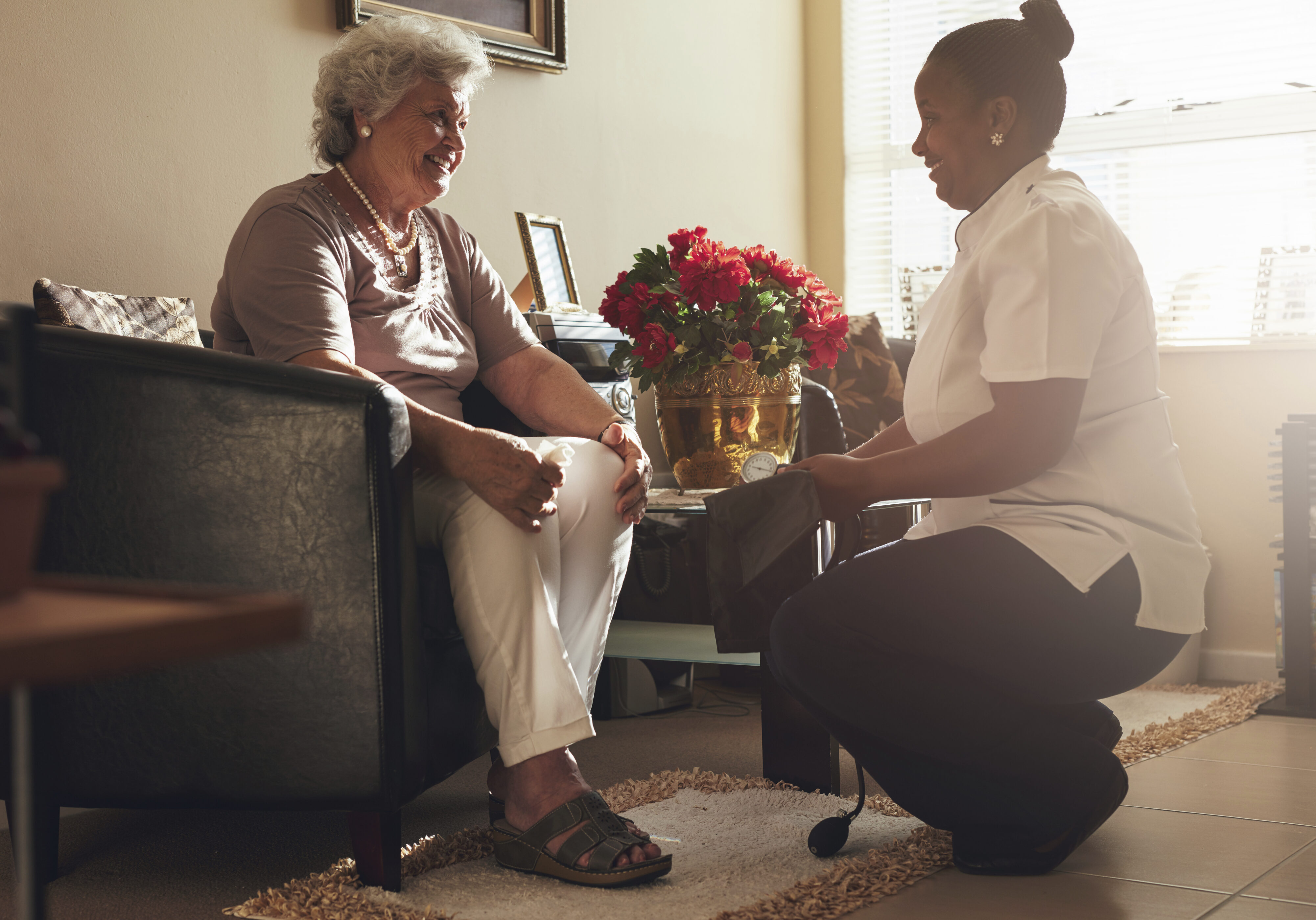 Senior woman sitting on a chair at home with female caregiver holding blood pressure gauge.  Female nurse visiting senior patient for checking blood pressure.